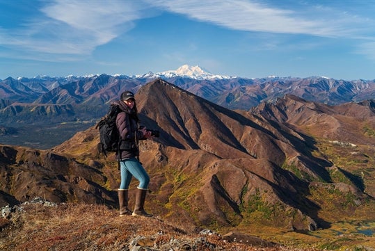 Photographer strikes a pose in front of an Alaskan mountain range, blue skies and wispy clouds