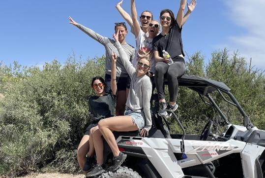 A group of young women sitting on the back of a white ATV smiling with their arms in the air and the desert behind them on a sunny day.