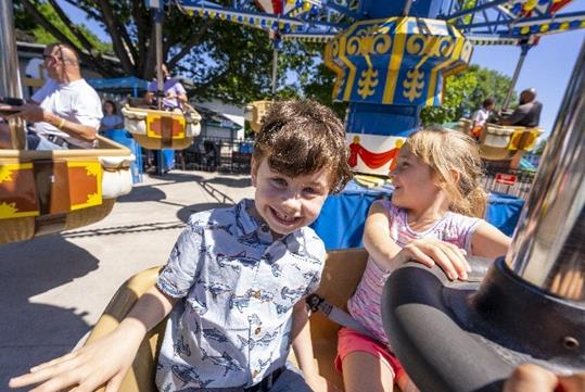 A young girl and boy smiling as they ride a carousel together, with other park guests enjoying the ride in the background on a sunny day.