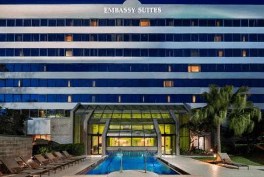 A small bright blue outdoor swimming pool with lounge chairs on the left and a palm tree on the right with the Embassy Suites behind it.