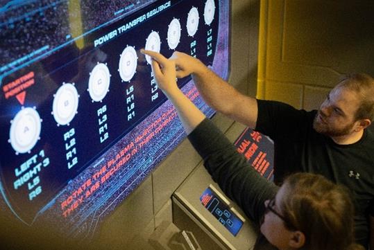 Two players pointing up at the Power Transfer Sequence chart mounted to the wall in front of them in the Star Trek Escape Room.