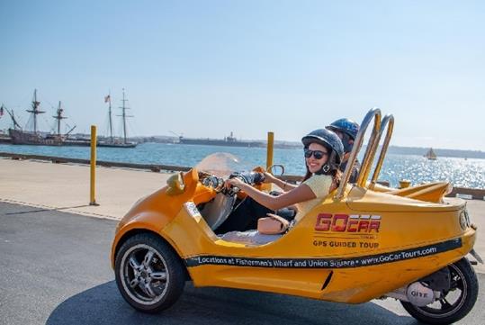 Tourists driving a yellow GoCar on a sunny day with water and ships in the background on the Experience San Diego GoCar Tour.