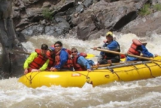 A group of people in red life jackets in a yellow raft splashing through the water with rocks behind them.