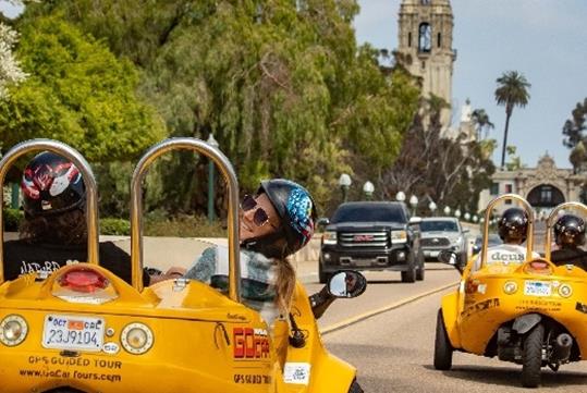 Two yellow GoCars with passengers wearing helmets driving them down a busy road in San Diego with palm trees to their right.