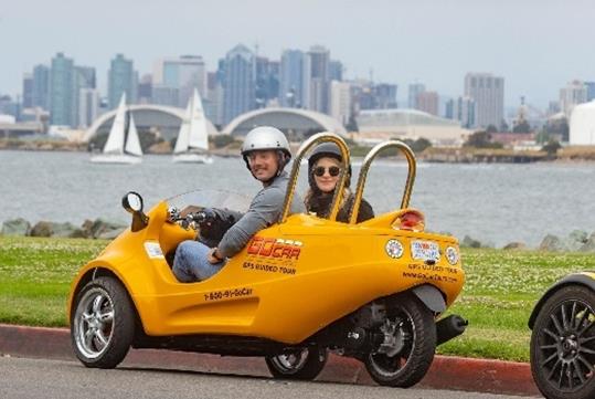 Tourists wearing helmets sitting in a yellow GoCar parked on the side of the road with boat in the water and the city in the background.