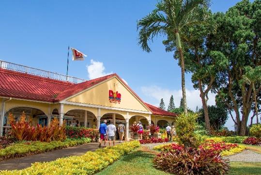 The front entrance to the Dole Pineapple Plantation with beautiful tropical landscaping and people walking into the building.