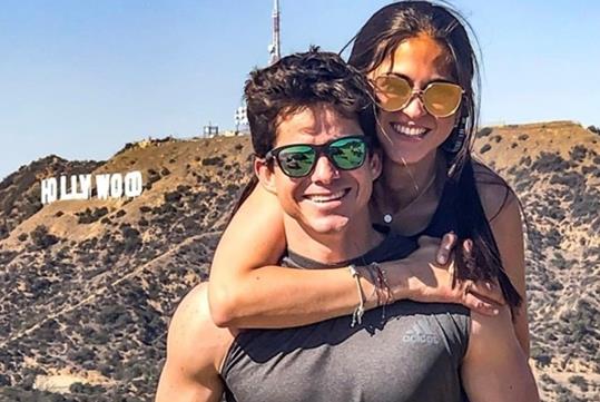 A man and woman wearing sunglasses with the woman on the mans back smiling with the Hollywood Sign in the background.