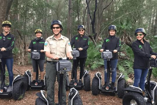 Group of tourists wearing black jackets on segways in the forrest with their tour guide wearing a khaki shirt on a segway in front of them.
