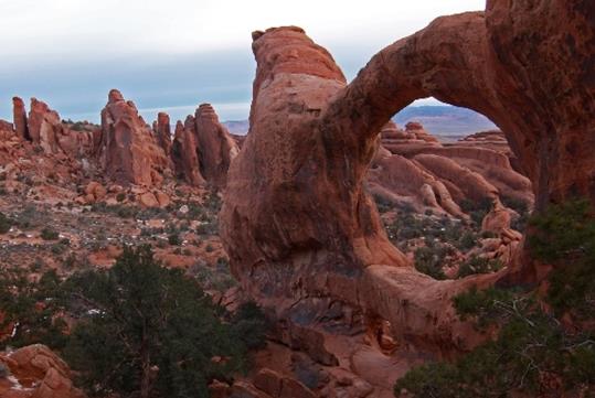 Stunning single arch rock formation at the Arches National Park with foliage scattered everywhere and a cloudy sky overhead.