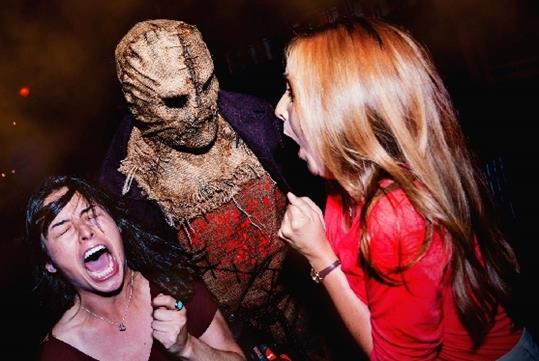 Two women with their mouths open screaming, one with her eyes shut, at a performer in a dirty burlap mask with no eyes.