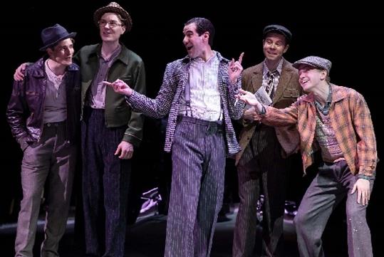 A group of men in 1920s style clothing gathered together talking and laughing on stage in the musical Harmony.