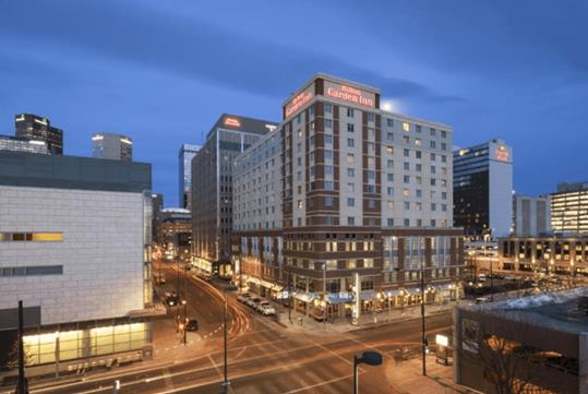 Wide shot of the exterior of the Hilton Garden Inn Denver Downtown on a cloudy night with cars zooming by.