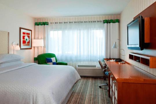 A hotel room with a king bed, a large window letting sun in, a green accent chair, a desk, a dresser, and a TV mounted to the wall.