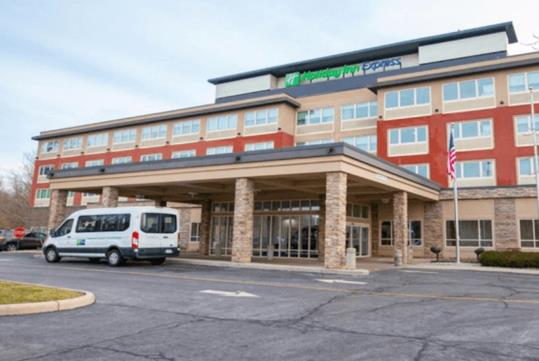 The tan stone and red exterior of the Holiday Inn Express Columbus Airport – Easton with the hotel shuttle parked in front.