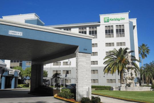 The front covered entrance and driveway of the Holiday Inn Orlando International Dr-ICON with palm trees and a fountain to the right.