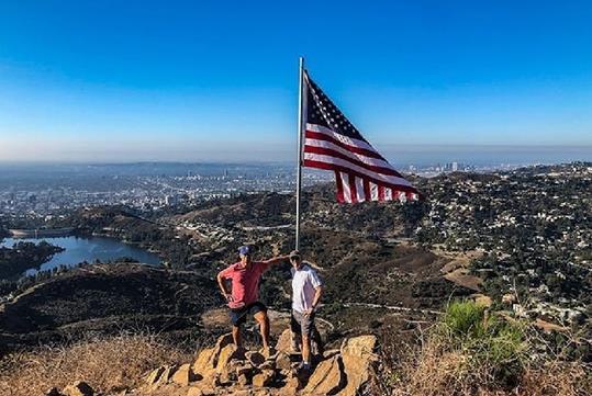 Two men in shorts and tee shirts standing next to an American Flag blowing in the wind on the Hollywood Sign Hike.