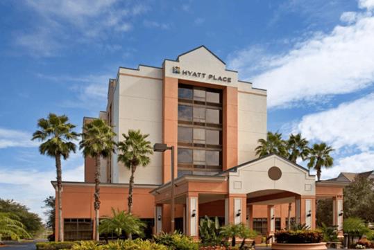 The front exterior of the Hyatt Place Orlando with several palm trees in front of it and a blue cloudy sky overhead.