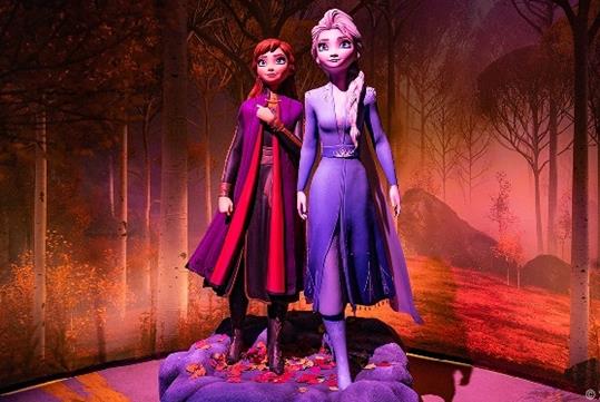 Two life-sized figures of Anna and Elsa from Disney's "Frozen" stand together on a rocky mound with autumn leaves, with a fall forest backdrop.