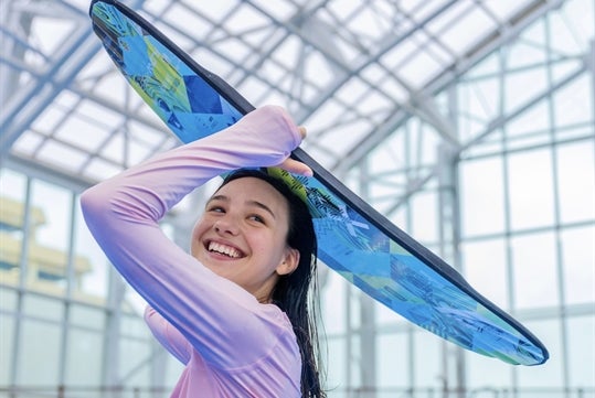 A girl in a pink long sleeve swim shirt smiling while holding a blue wake board over her head.