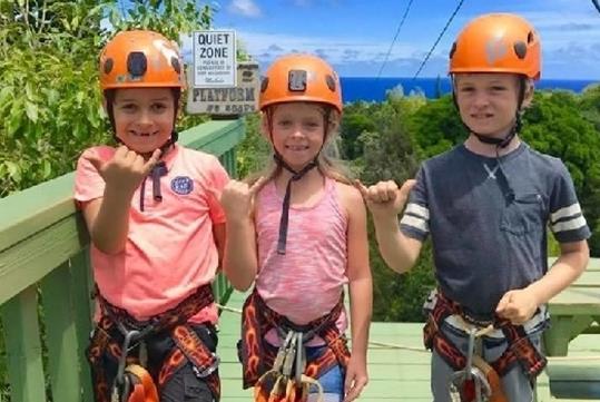 Three young kids in orange helmets and zip lining harness with their hangs doing the shaka sign on a sunny day.