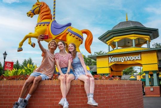 Three teenagers snapping a selfie while seated on a brick wall next to a horse carousel figure at the entrance to Kennywood.