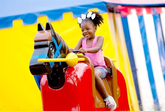 A smiling young girl riding a colorful, toy horse in a playful setting with bright, cheerful backgrounds.