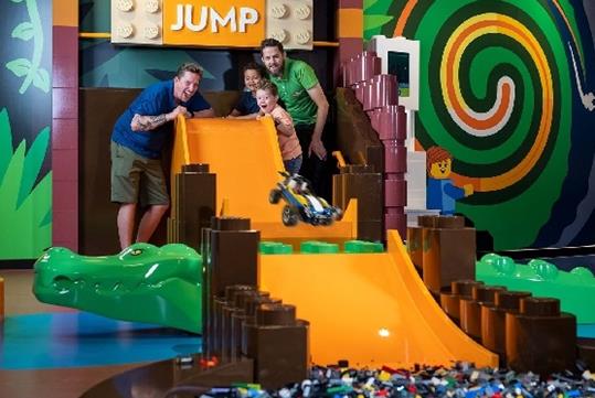 Two parents and two children at the top of an orange ramp with a giant LEGO alligator in the middle and a toy car in the air.