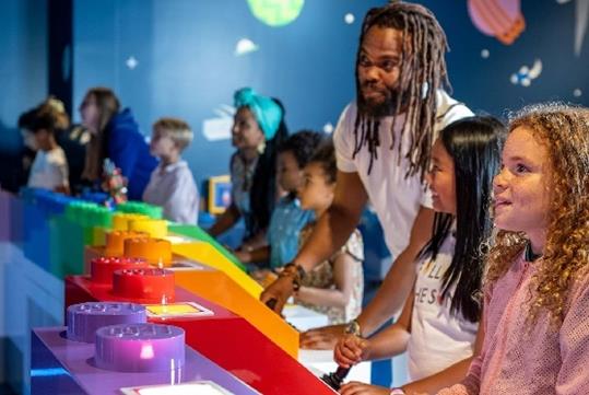 A line of several parents and children playing games and smiling at LEGO Discovery Center Boston.
