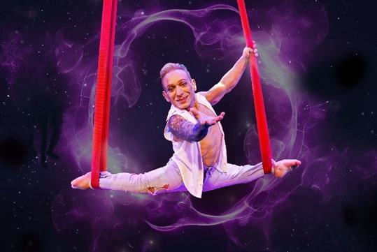 Alan Silva doing the splits with one of his arms out with each of his feet in red aerial silks at Le Grand Cirque Presents Adrenaline.