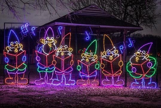 A lighting display of Christmas gnomes singing with music notes above their heads at night at the Lights of Joy Christmas Drive-Thru.