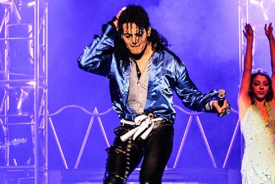 A Michael Jackson impersonator in a shiny blue shirt with back up dancers on either side of him on stage.