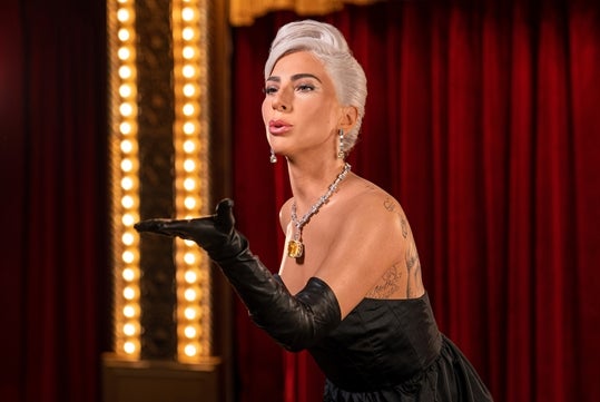 A wax figure of Lady Gaga, dressed in an elegant black gown, standing on a stage with a rich red curtain and ornate lighting at Madame Tussauds Hollywood.