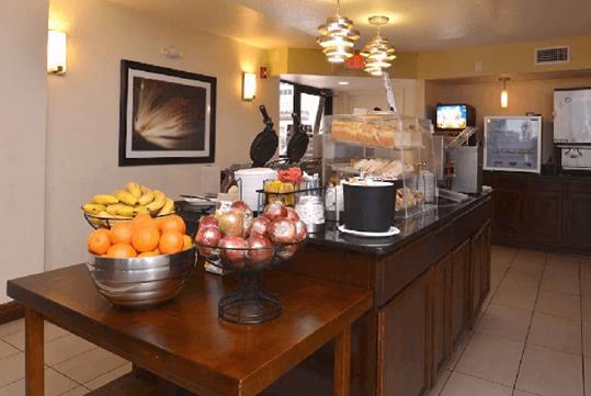 A breakfast buffet area with assorted fresh fruits and other breakfast items on a dark brown island with drink machines in the background.