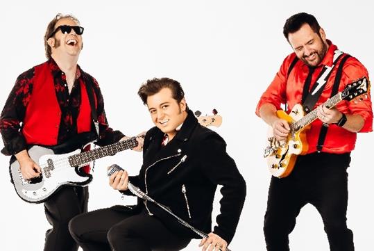 Two men in red and black rocker shirts playing guitars with a man in all black squatting with a microphone stand in between them