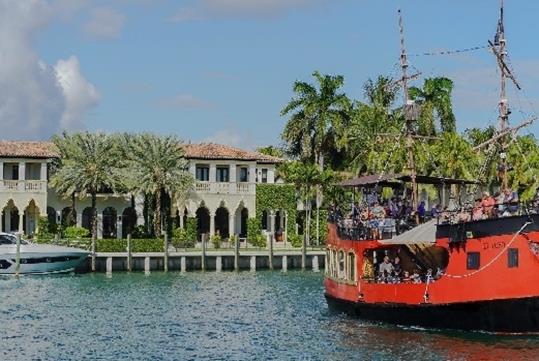 A red & black Pirate Ship in blue water cruising in front of a stunning mansion with palm trees surrounding it on a sunny day in Miami.