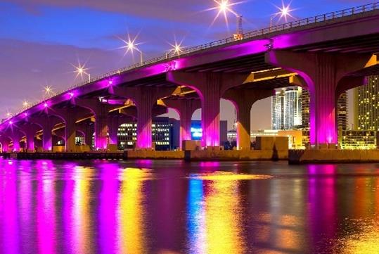 View of the underside of a bridge over water at night with purple lights on the side and street lamps on top, both colors of light reflect on the water below.