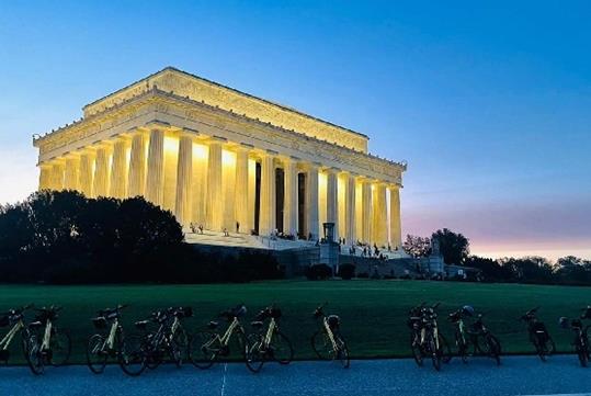 A row of bikes lined up near the grass in front of the Lincoln Memorial with tourists walking up the steps at dusk.