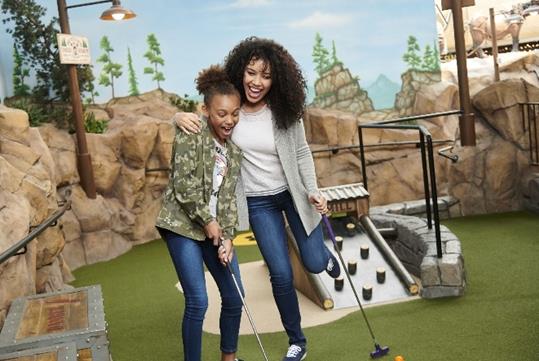 A young and middle age women smiling and laughing while playing mini golf with a wall of rock behind them at Moose Mountain Adventure Golf at Mall of America.