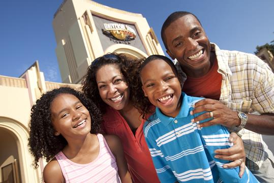 A family of four posing together for a photo with the entrance to Universal Studios Orlando in the background on a sunny day.