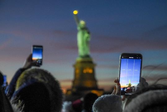 Tourists in winter clothing holding their phones in the air to get a photo of Lady Liberty at dusk.
