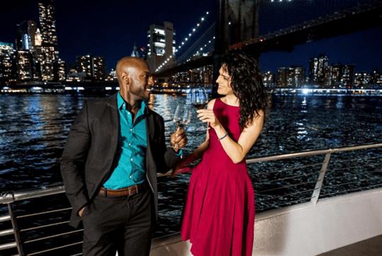 A man and woman talking and cheering their wine glasses together near the railing of a cruise boat with NYC in the background at night.