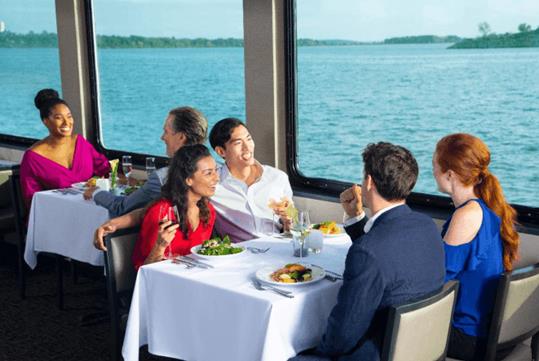 Guests chatting and sitting down for dinner with large windows next to them on the New York Signature Lunch Cruise.