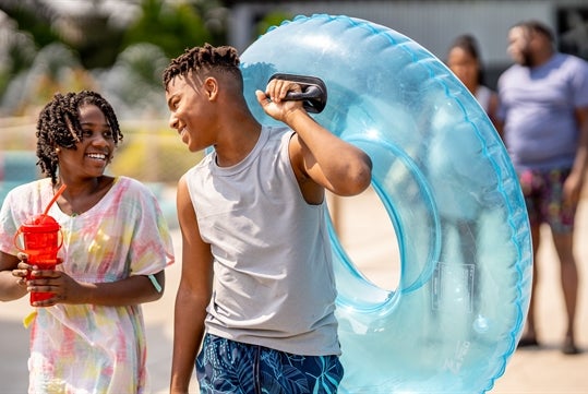 Two boys enjoy a day at the water park, one wearing a white t-shirt adorned with colorful paint-like splashes and dark shorts, holding a red cup, while the other wears a gray tank top and blue swim shorts, carrying a large blue inflatable ring, captured against a backdrop of blurred park goers at Noah's Ark in Wisconsin Dells, Wisconsin.