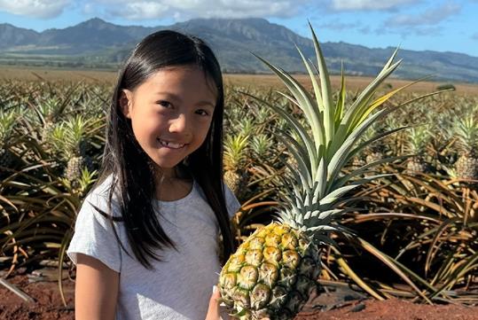 A young girl with dark hair smiling and holding a ripe pineapple in her hands with a field of pineapple behind her.