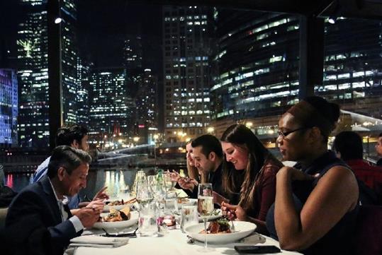 A group of friends seated at a white table enjoying their dinner with a large window showing the city of Chicago at night behind them.