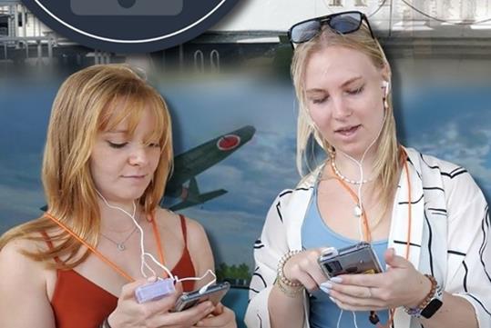 Two women holding smartphones and earbuds while on the USS Arizona Narrated Multimedia Tours with a poster behind them.