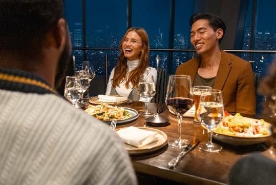 A group of friends gathered around a table enjoying dinner and drinks at the One World Observatory with the windows behind them showing NYC.
