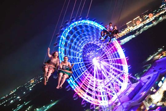 Guests laughing and screaming while riding the Orlando Starflyer at ICON Park at night with a glowing ferris wheel behind them.