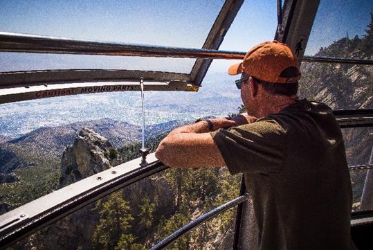 A man in an orange baseball cap inside the tramcar leaning on the railing of an open window looking at the mountains at Palm Springs Aerial Tramway.