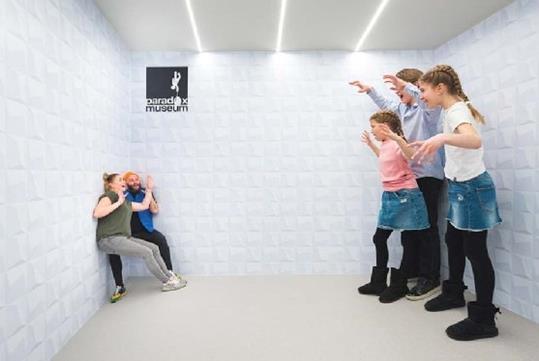 An optical illusion room with small looking cowering parents on one side and three giant children with their hands up on the other.